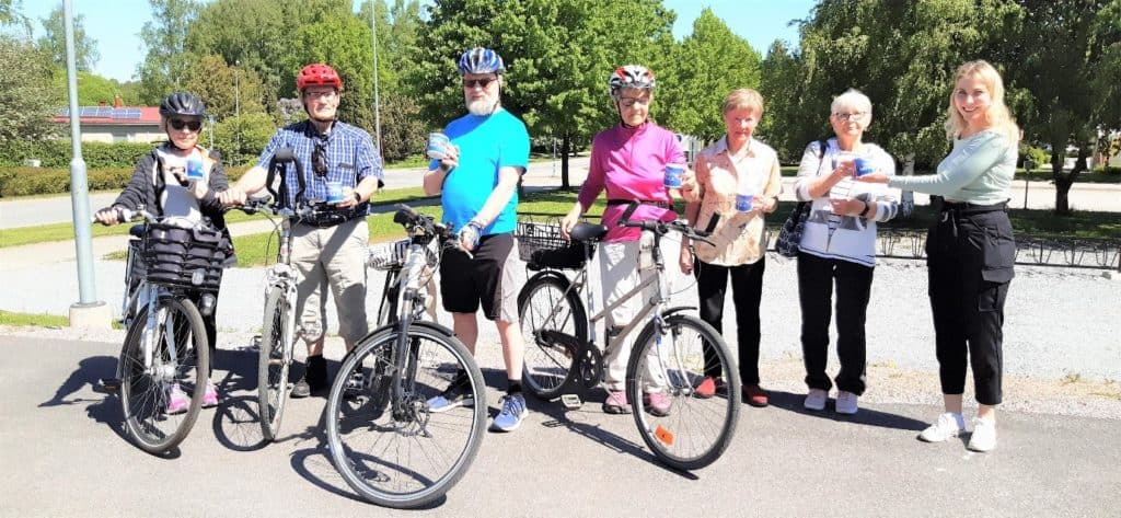 Kuvassa on kuusi henkilöä, joilla on Team Hattula -mukit kädessä. Neljällä henkilöllä on myös polkupyörät ja pyöräilykypärät. Vihreäpaitainen nainen on ojentamassa voittomukia. 